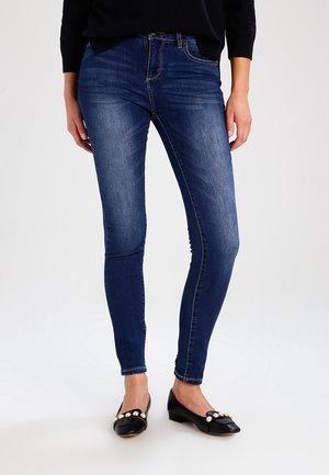 Vrouw die donkerblauwe skinny jeans en zwarte platte schoenen met parelversieringen draagt, staand tegen een effen lichte achtergrond.