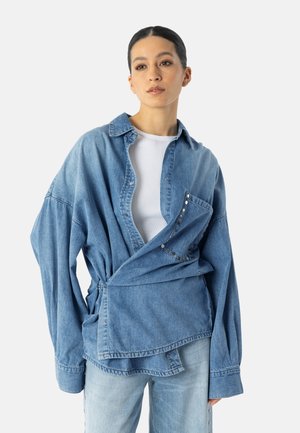 Kvinde med mørkt hår samlet bagud iført oversize blå denimjakke med nittet lomme over hvid skjorte og lyseblå jeans, neutral baggrund.