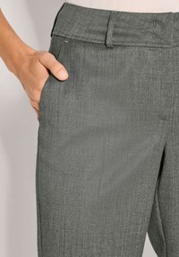 Close-up van een hand in de voorzak van grijze, op maat gemaakte broek met riemlussen en subtiele textuur.