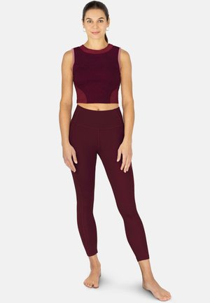 Frau steht barfuß, trägt ein kastanienbraunes ärmelloses Crop-Top und passende hoch taillierte Leggings vor einem einfarbigen hellen Hintergrund.