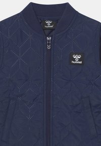 Marinblå vadderad bomberjacka med diamantmönster, dragkedjestängning, ribbad krage och en liten logotypapplikation på bröstet. Texturerad tyg.