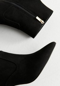 Stivali ankle in suede neri con punta appuntita e cerniera laterale. Presentano una texture liscia e cuciture sottili. Dettaglio in metallo dorato della cerniera.