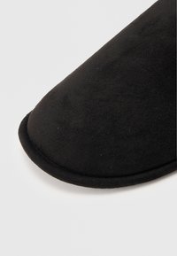 Zapatilla negra con punta redondeada, hecha de un suave tejido cepillado. Textura suave y detalles de costura alrededor del borde.