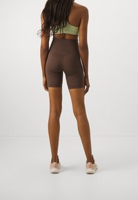 Hnědé šortky s vysokým pasem z pružné látky, s těsným střihem a minimálním šitím, doplněné světle růžovými sportovními botami.