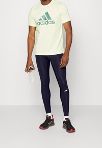 Ljust gult t-shirt med kort ärm och grön Adidas-logga, marinblå leggings och svarta träningsskor med röda detaljer. Vattenflaska i handen.