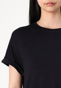 T-shirt noir à manches courtes en tissu doux et lisse. Design à col ras du cou avec des manches roulées, mettant en avant une silhouette minimaliste et classique.