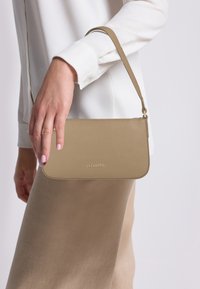 Donna che tiene una piccola borsa in pelle beige con manico corto, indossando una blusa bianca e una gonna beige.