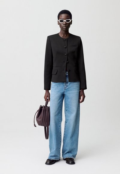 Mujer con gafas de sol redondas blancas, blazer negro abotonado, jeans de pierna ancha azul claro, zapatos negros y sosteniendo un bolso de cuero marrón tipo cubo.