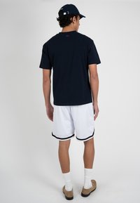 Hombre de espaldas con gorra negra, camiseta azul marino, pantalones cortos de malla blanca con ribete negro, calcetines blancos y zapatos marrones sin cordones.