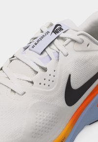 Bela tekaška športna obuvala Nike s črnim logotipom, belimi vezalkami ter oranžnim in modrim podplatom, približan zgornji del z napisom "Run Structure 25".