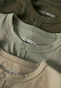 Kolm puuvillast särki erinevates vaiksetes rohelise ja beeži toonides, ümara kaelusega ja nupudega, millel on silt "TWO SOON."