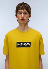 Gul kortærmet t-shirt lavet af bomuld, med et sort rektangulært logo med hvid tekst på forsiden. Glat tekstur, rund halsudskæring.