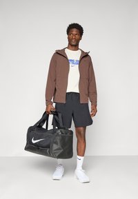 Mand i brun jakke, hvid grafisk t-shirt, sorte shorts, hvide sneakers og høje sokker, der holder en stor sort Nike duffel taske.