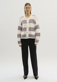 Cardigan a righe bianche e grigie con una texture soffice, dotato di chiusura a bottoni e tasche frontali, abbinato a pantaloni neri e scarpe a punta.