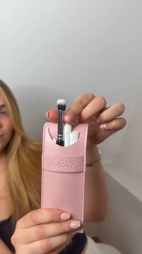 Hånd holder pink ZOEVA pung, der indeholder to makeupbørster med hvide børster, mod en ensartet lys baggrund.
