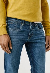 Jean en denim bleu à coupe droite, avec une légère décoloration, cinq poches et un petit logo sur la poche arrière. Associé à un pull jaune.