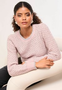 Világos rózsaszín pulóver textúrázott gyémántmintával, fényes részletekkel a nyakrészen és az ujjak végén, valamint bordázott szegélyekkel. A modell egy semleges felületen ül.