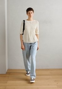 Maglione lavorato a maglia di colore crema con maniche corte, jeans azzurri morbidi e scarpe da ginnastica marroni con motivo, completati da una borsa a spalla nera.