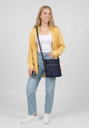 Junge Frau in gelbem Hemd, weißem Tanktop, blauen Jeans und weißen Sneakers, die ein Smartphone und eine marineblaue Umhängetasche hält, lächelnd und nach rechts schaut.