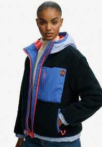 Vrouw in een zwart-blauwe fleecejack met een rode rits en capuchon, staand met één hand in de zak tegen een witte achtergrond.