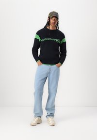 Pulover tricotat de culoare bleumarin închis, cu accente verzi și logo, purtat cu jeansi de culoare albastru deschis și adidași bej, completat cu o șapcă cu model camuflat.