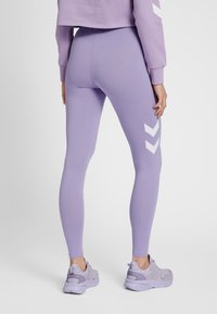 Lavendelkleurige leggings, slank model, gladde textuur, met witte chevron accenten aan de linkerkant. Gecombineerd met lichtpaarse sneakers.