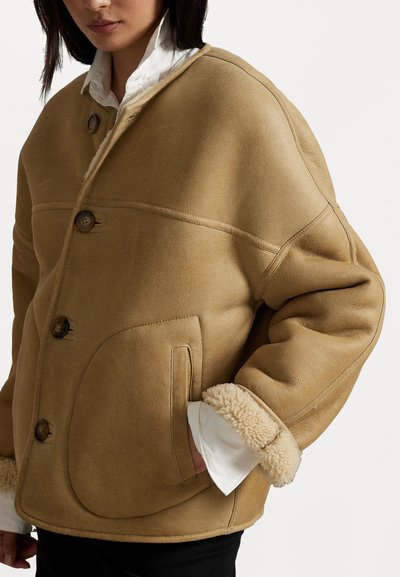 Giacca in shearling color tan con vestibilità rilassata, grandi tasche, tre bottoni e texture morbida, dotata di polsini in pelliccia a contrasto.