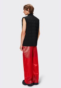 Gilet noir matelassé sans manches avec un col haut, associé à un pantalon rouge brillant et ample, présentant une texture lisse et une finition brillante.