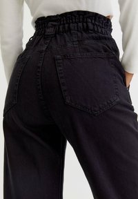 Vrouw in zwart, hooggewrichte broek met elastische tailleband en achterzakken, gecombineerd met een witte blouse met lange mouwen.