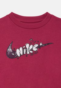 Džemperis tamsiai raudonas, su ryškiu juodai baltu Nike logotipo grafiniu atvaizdu, papuoštu žvaigždėmis ir širdimis. Raišteliai prie kaklo.