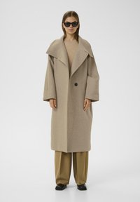 Cappotto in lana beige con ampio colletto, maniche oversize, chiusura a un bottone, tasche laterali e design dritto. Occhiali da sole neri indossati.