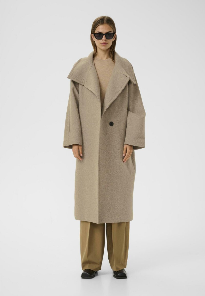 Cappotto in lana beige con ampio colletto, maniche oversize, chiusura a un bottone, tasche laterali e design dritto. Occhiali da sole neri indossati.