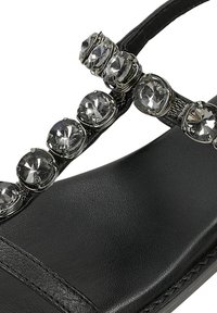 Sandal nero con una fascia testurizzata adornata da numerosi strass trasparenti e dettagli metallici sulla parte superiore. Design con suola piatta.