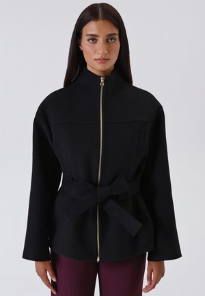 Giacca nera con zip realizzata in tessuto morbido, caratterizzata da un alto colletto, tasche laterali e una cintura in vita legata. I dettagli in zip dorata aggiungono un tocco di stile.