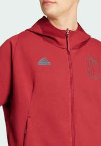 Red zip-up hoodie made of soft fabric, features a gray Adidas logo and embroidered crest. Includes a hood and side pockets.