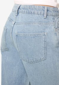 Šviesiai mėlyni džinsai iš džinso audinio, dėvimi asmens, matomos nugaros kišenės ir diržo kilpos, su matoma balta juosmens juosta virš džinsų.