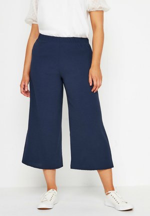 Kobieta w granatowych szerokich spodniach o długości do połowy łydki, białej bluzce z krótkim rękawem i białych trampkach, stojąca na tle jednolitego białego tła.