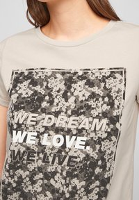 Světle béžové tričko s květinovým potiskem a černobílým textem, který říká "SNÍME. MILUJEME. ŽIJEME." v obdélníkovém designu.
