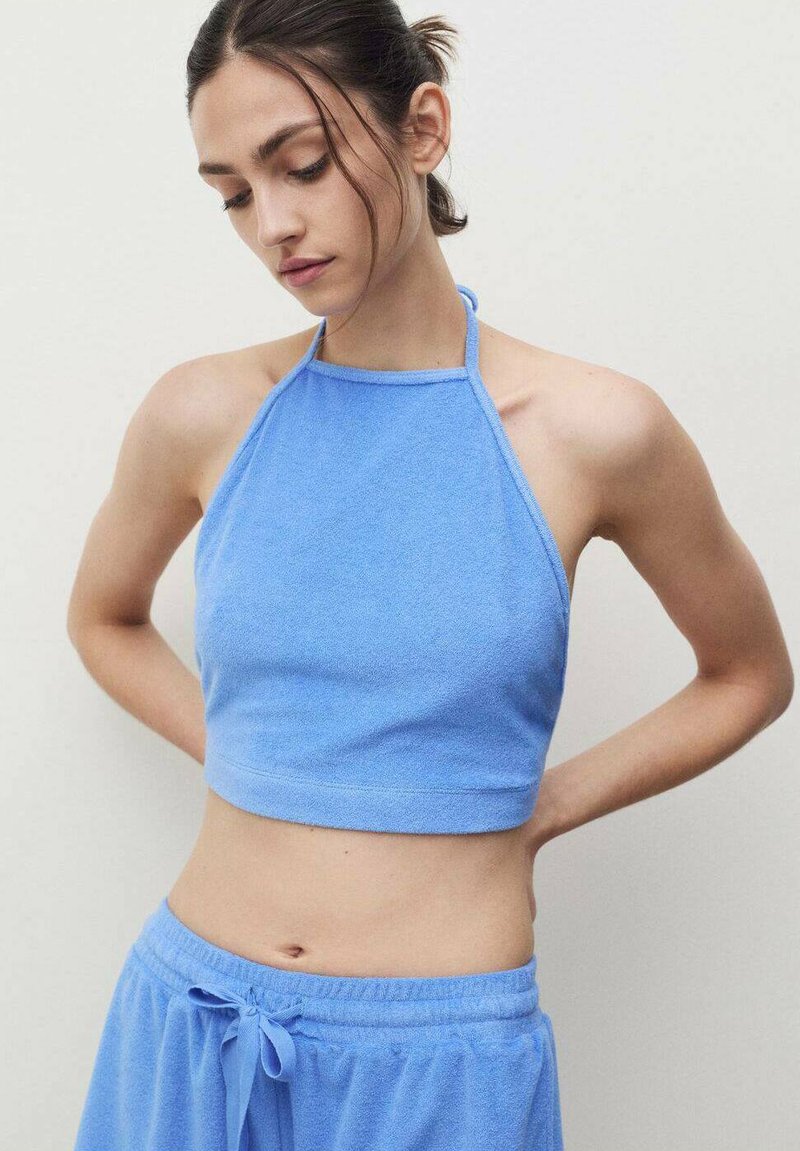 Modrý halter cropped top z froté látky se tenkými ramínky, jemnou texturou a přiléhavým střihem. V kombinaci s odpovídajícími modrými volnými šortkami.