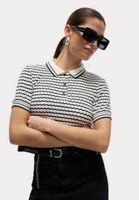 Poloshirt met kraag in een crèmekleurige basis met marineblauwe golvende strepen. Korte mouwen met marineblauwe boorden. Zwarte riem met een zilveren gesp, gecombineerd met een donkere rok.