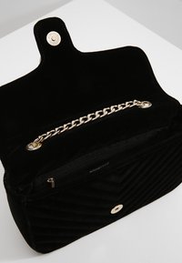 Svart vadderad sammetshandväska med kedjedetalj, knappstängning och ett dragkedjefack. Har en slät innersida.