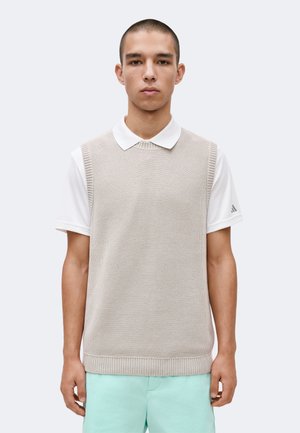 Young man wearing a beige knitted vest over a white polo and light blue shorts, standing against a plain light background.