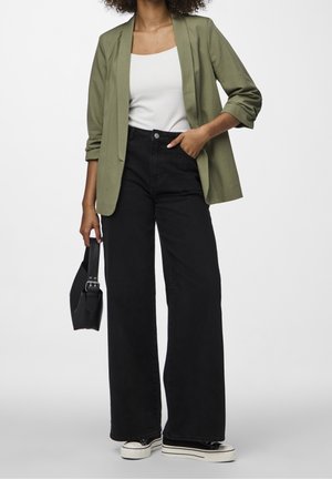 Blazer vert olive, débardeur blanc, jeans noirs à taille haute et jambes larges, et sac à main noir. Baskets avec semelles blanches et lacets noirs.