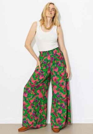 Femme blonde debout contre un mur blanc, portant un haut sans manches blanc, un pantalon vert à jambes larges avec un imprimé floral rose et violet, et des chaussures tissées marron.