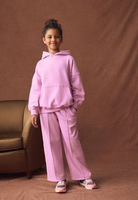 Felpa rosa con cappuccio e ampia tasca frontale, abbinata a pantaloni a gamba larga coordinati. Tessuto liscio con vestibilità comoda. Sneakers con un motivo vivace.