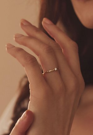 Gouden ring met een gladde band en een enkele fonkelende steen, gedragen op een hand met bleke, gemanicuurde nagels tegen een zachte, neutrale achtergrond.
