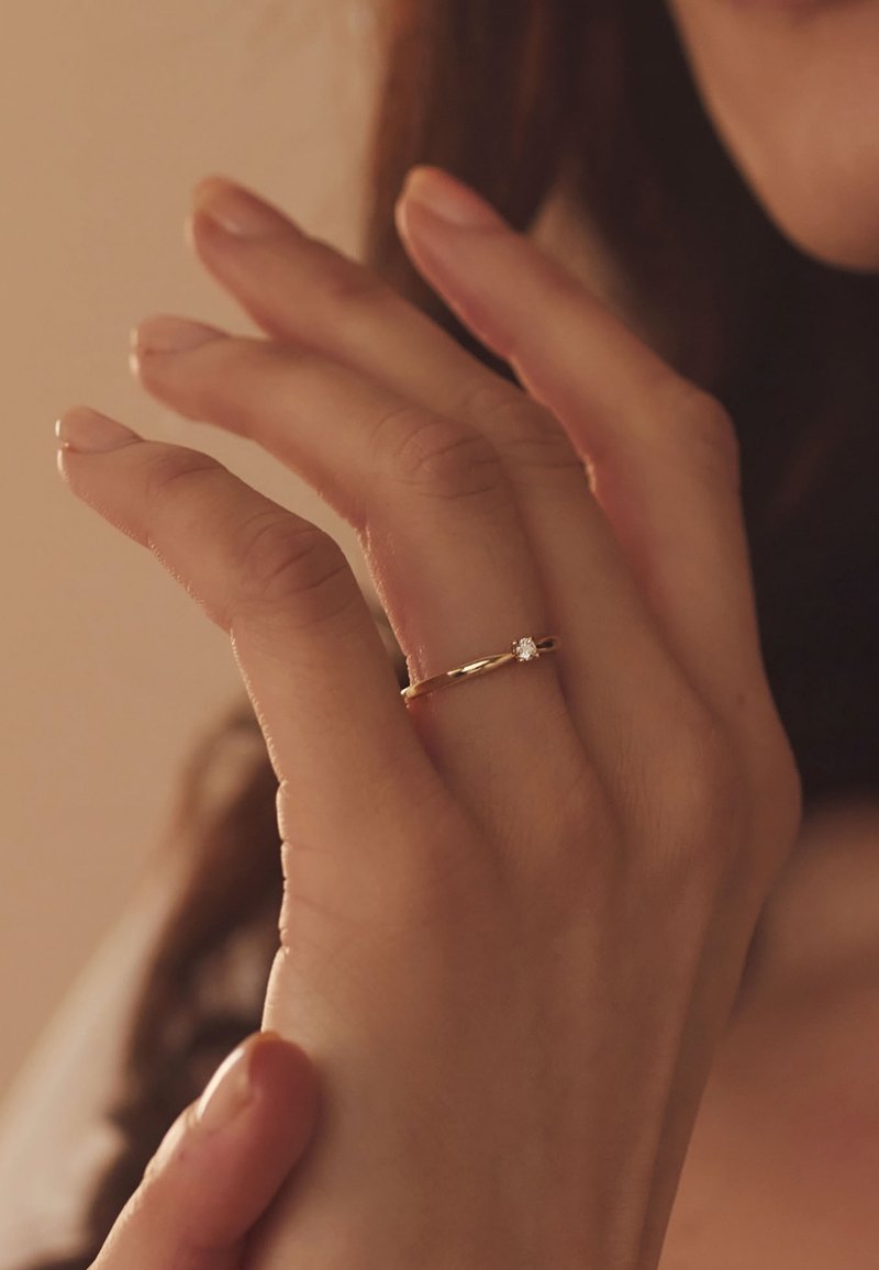 Gouden ring met een gladde band en een enkele fonkelende steen, gedragen op een hand met bleke, gemanicuurde nagels tegen een zachte, neutrale achtergrond.