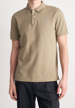 Homme portant un polo beige à manches courtes avec deux boutons, associé à un pantalon noir sur un fond uni.