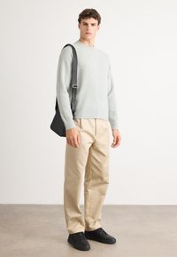 Homme portant un pull gris clair, un pantalon beige, des baskets noires, portant un sac à bandoulière noir sur fond blanc plain.
