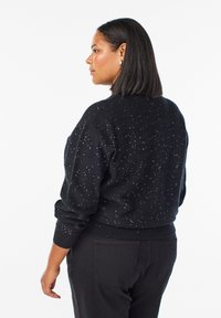 Černý, texturovaný svetr s kratší siluetou, zdobený malými stříbrnými třpyty, v kombinaci s černými kalhotami.